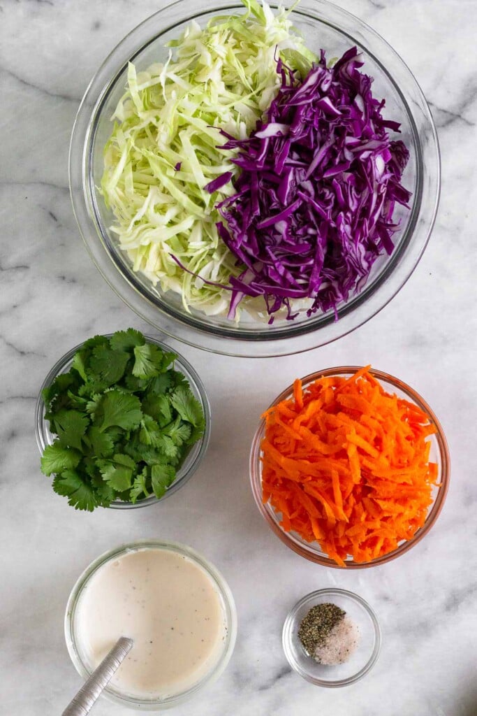 White marble counter with a bowl of shredded green and red cabbage, a bowl of shredded carrots, a bowl of salt and pepper, a jar of yogurt dressing, and a bowl of cilantro.