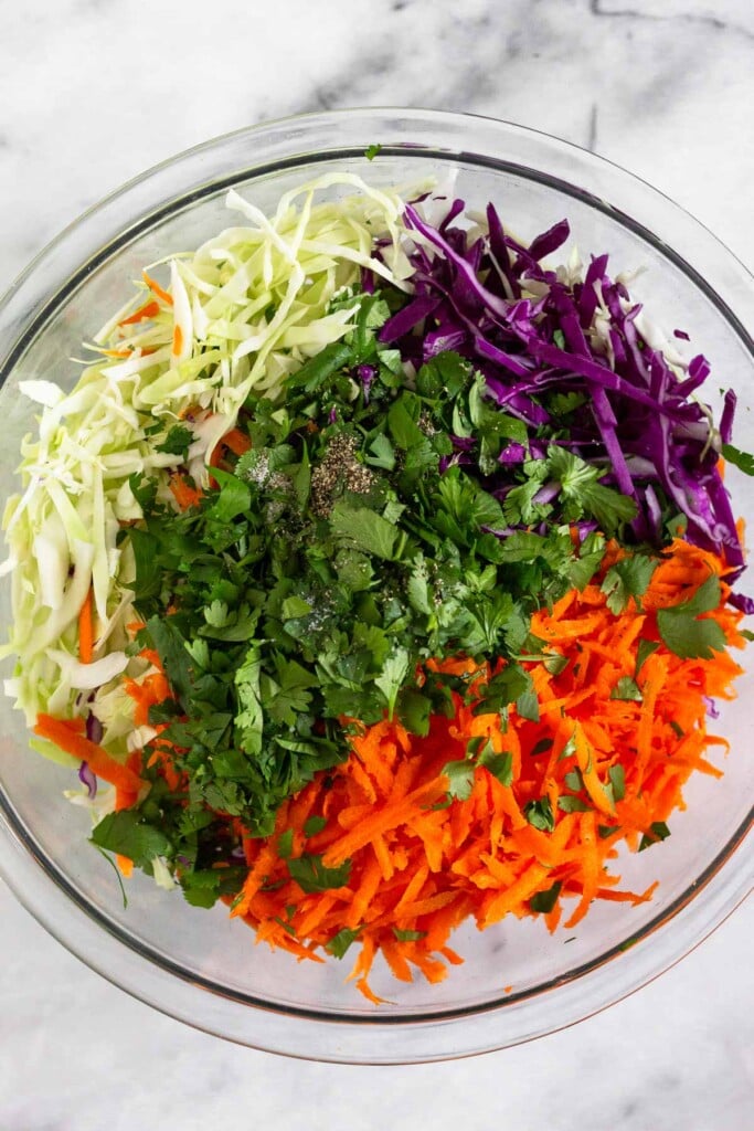 Large glass bowl filled with shredded green and red cabbage, shredded carrots, cilantro, salt, and pepper.