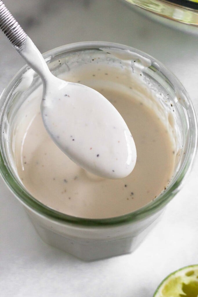 A jar of healthy coleslaw dressing with a spoon full of dressing overtop of it. In front of the jar is half a juiced lime.