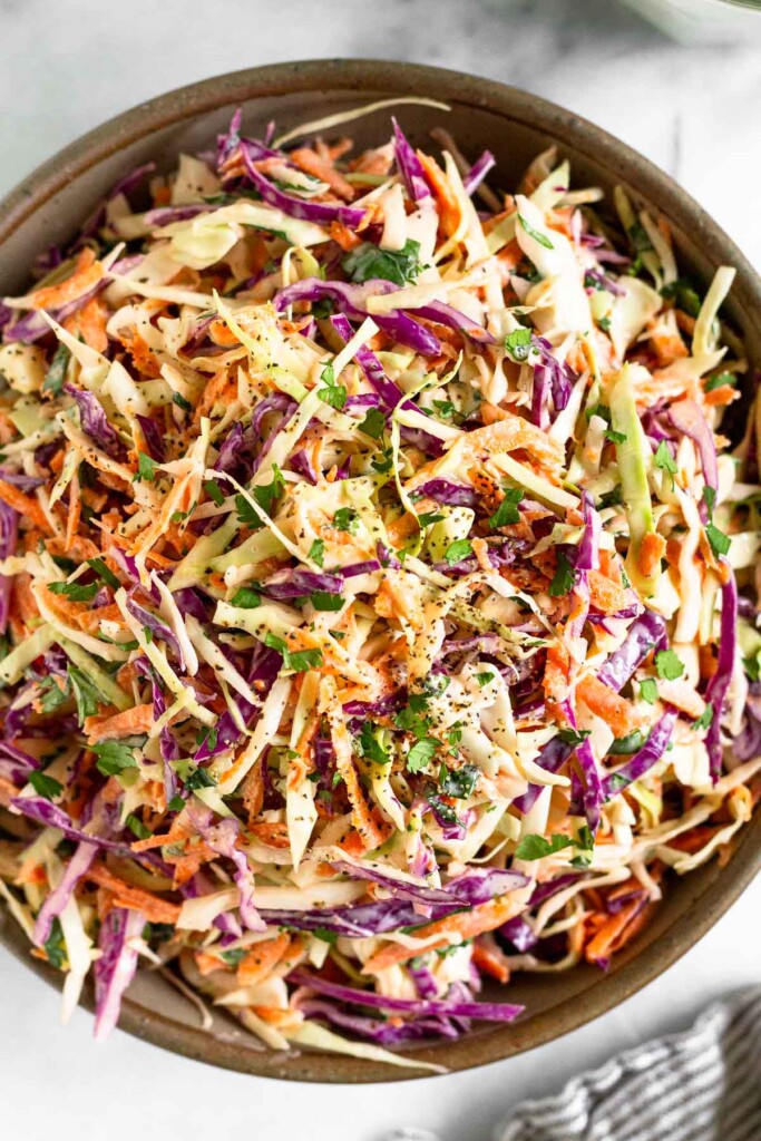 Overhead shot of coleslaw with greek yogurt garnished with fresh cilantro and black pepper in a large bowl. A striped towel is off to the side of the bowl.