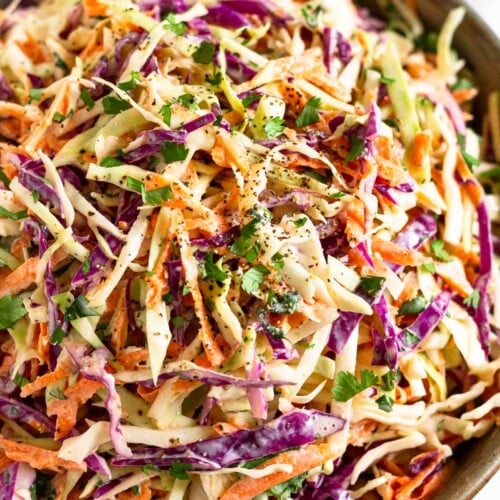 A closeup of a large bowl of healthy coleslaw garnished with fresh cilantro and black pepper. A striped towel is in front of the bowl.