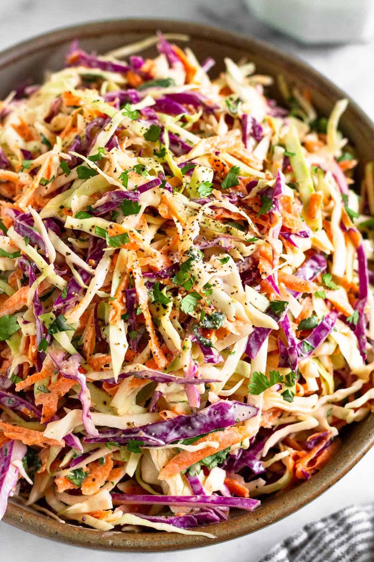 A closeup of a large bowl of healthy coleslaw garnished with fresh cilantro and black pepper. A striped towel is in front of the bowl.