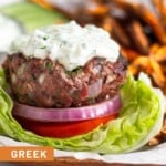 Lamb burger sitting on a lettuce cup with sliced tomato, red onion, and topped with tzatziki sauce. Behind the burger are sweet potatoes fries, sliced cucumber, and a dish of tzatziki sauce.