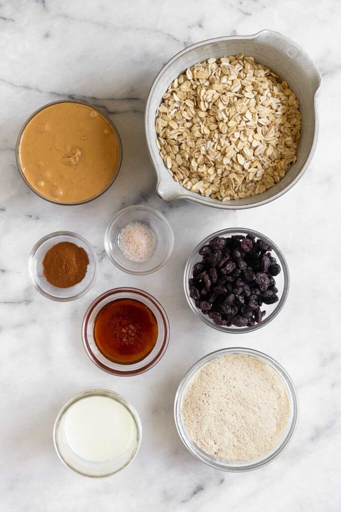 White marble counter with a bowl of rolled oats, a bowl of raisins, a bowl of vanilla protein powder, a jar of milk, a bowl of honey, a bowl of cinnamon, a bowl of salt, and a bowl of cashew butter.