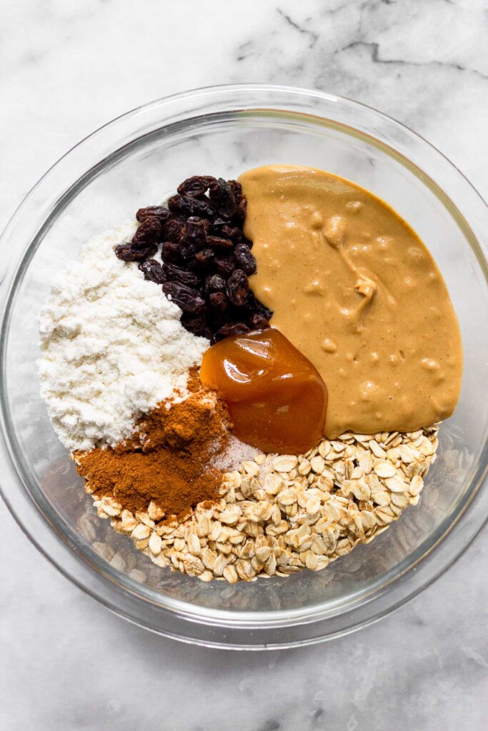 Glass bowl filled with rolled oats, protein powder, raisins, cashew butter, honey, cinnamon, and salt.