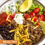 Taco bowl filled with ground beef taco meat, black beans, salsa, cilantro lime rice, sliced avocado, lettuce, diced tomatoes, shredded cheese, sour cream, and a lime wedge garnished with cilantro. In front of the bowl is another taco bowl and behind it is a dish of cilantro and bowl of tortilla chips.