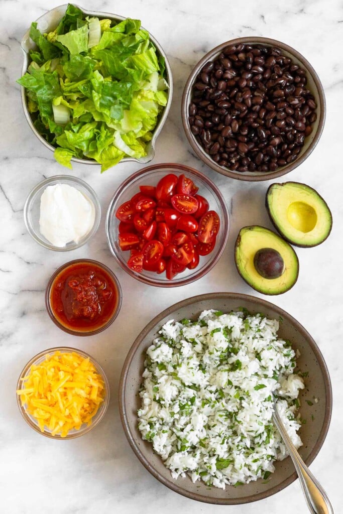 White marble counter with a bowl of black beans, an avocado that has been cut in half, a bowl of cilantro lime rice, a bowl of shredded cheese, a bowl of salsa, a bowl of sour cream, and a bowl of chopped lettuce.