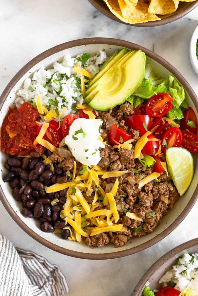 A large white bowl filled with ground beef taco meat, black beans, salsa, cilantro lime rice, sliced avocado, lettuce, tomatoes, shredded cheese, sour cream, and a lime wedge. It is garnished with cilantro. Around the bowl is a striped towel, another taco bowl, and a bowl of tortilla chips.