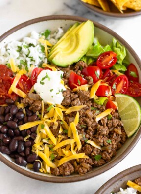 Taco bowl filled with ground beef taco meat, black beans, salsa, cilantro lime rice, sliced avocado, lettuce, diced tomatoes, shredded cheese, sour cream, and a lime wedge garnished with cilantro. In front of the bowl is another taco bowl and behind it is a bowl of tortillas chips.