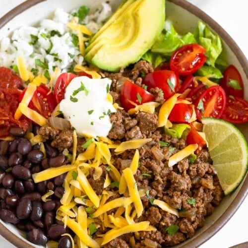 Taco bowl filled with ground beef taco meat, black beans, salsa, cilantro lime rice, sliced avocado, lettuce, diced tomatoes, shredded cheese, sour cream, and a lime wedge garnished with cilantro. In front of the bowl is another taco bowl and behind it is a bowl of tortillas chips.