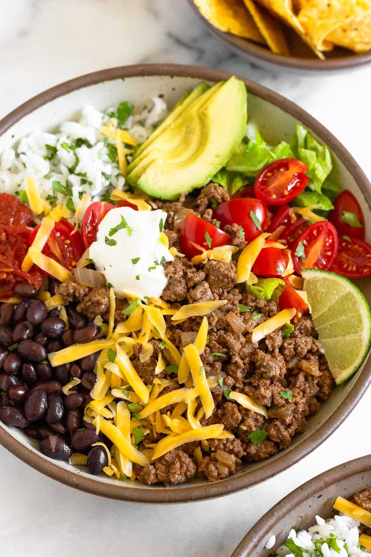 Taco bowl filled with ground beef taco meat, black beans, salsa, cilantro lime rice, sliced avocado, lettuce, diced tomatoes, shredded cheese, sour cream, and a lime wedge garnished with cilantro. In front of the bowl is another taco bowl and behind it is a bowl of tortillas chips.