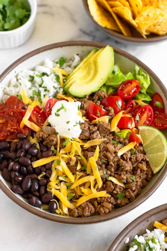 Ground beef taco bowl with black beans, salsa, cilantro lime rice, sliced avocado, lettuce, diced tomatoes, shredded cheese, sour cream, and a lime wedge. It is garnished with cilantro. In front of the bowl is another taco bowl and behind it is a dish of cilantro and bowl of tortilla chips.