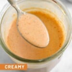 Creamy salsa dressing in a jar with a spoon full of dressing overtop of it. In front of the jar is half a lime and a teaspoon with spices in it. Behind the jar is a small dish of salsa.