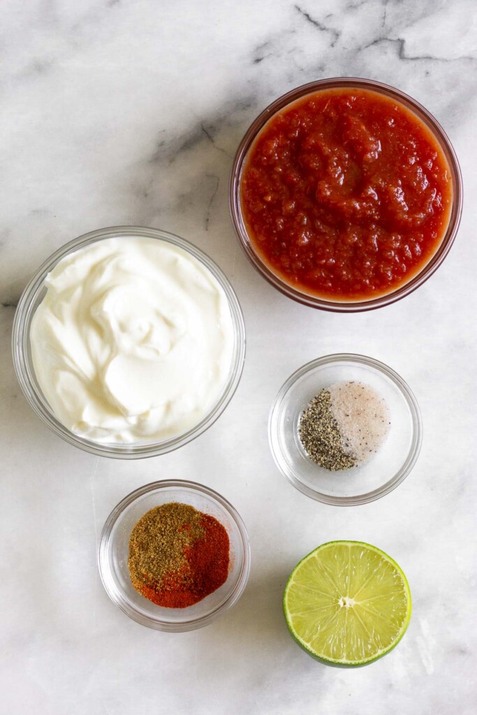 White marble countertop with a bowl of red salsa, a bowl of salt and pepper, half a lime, a bowl of spices, and a bowl of greek yogurt.