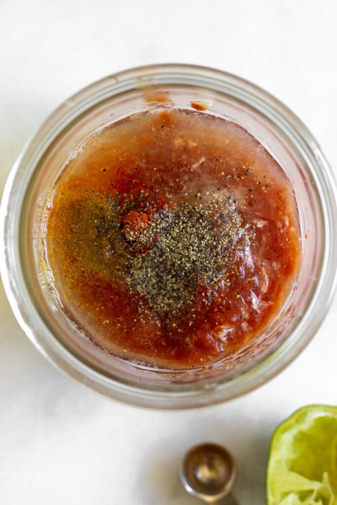 Overhead shot of a jar filled with yogurt, salsa, lime juice, and spices before it is mixed. Next to it is a half a juiced lime and an empty teaspoon.