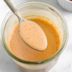 Creamy salsa dressing in a jar with a spoon full of dressing overtop of it. In front of the jar is half a lime and a teaspoon with spices in it. Behind the jar is a small dish of salsa.