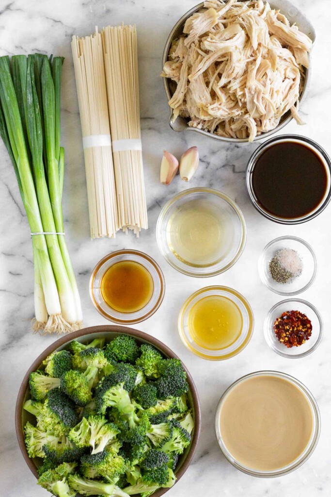 White marble counter with a bowl of cooked shredded chicken, a bowl of coconut aminos, a bowl of salt and pepper, an bow of red pepper flakes, a bowl of tahini, a bowl of broccoli florets, a bowl of honey, a bowl of vinegar, a bowl of sesame oil, a bunch of green onions, and 2 bunches of dry noodles.