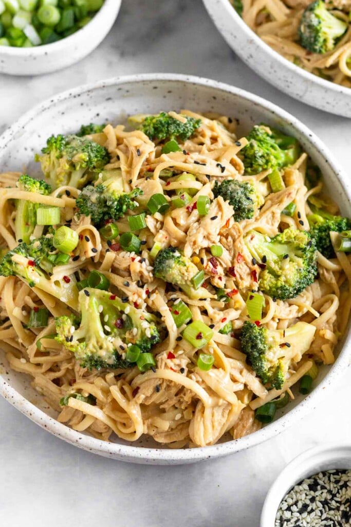A white bowl filled with sesame noodles with chicken and broccoli garnished with sesame seeds and red pepper flakes. Around the bowl is a dish of black and white sesame seeds, a dish of sliced green onions, and another bowl of sesame chicken noodles.