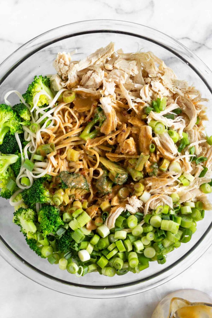 Large glass bowl with a cooked pasta, broccoli florets, and sliced green onion with a brown sauce poured on top of them. It is not mixed up yet.