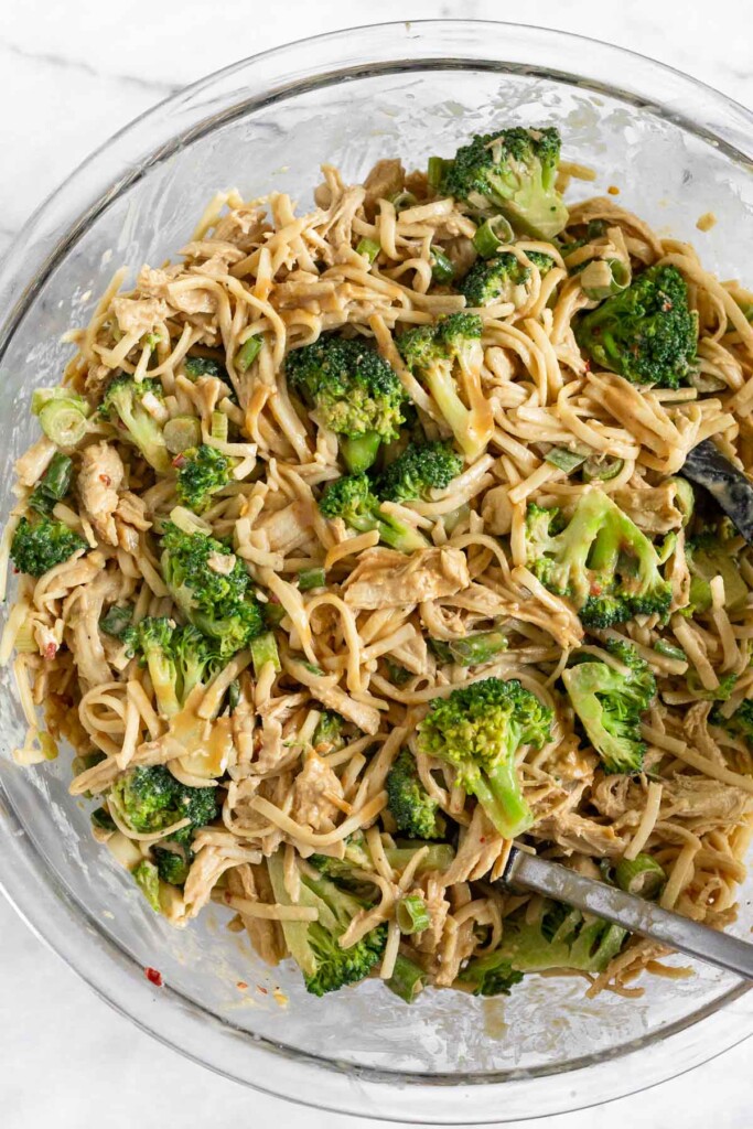 A large glass bowl filled with pasta, chicken, broccoli florets, and sliced scallions tossed in a creamy brown sauce.