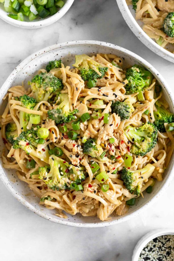 Sesame chicken noodles with broccoli and green onions in a white bowl. It is garnished with sesame seeds and red pepper flakes. Surrounding the bowl is a dish of black and white sesame seeds, a dish of sliced green onions, and another bowl of sesame chicken noodles.