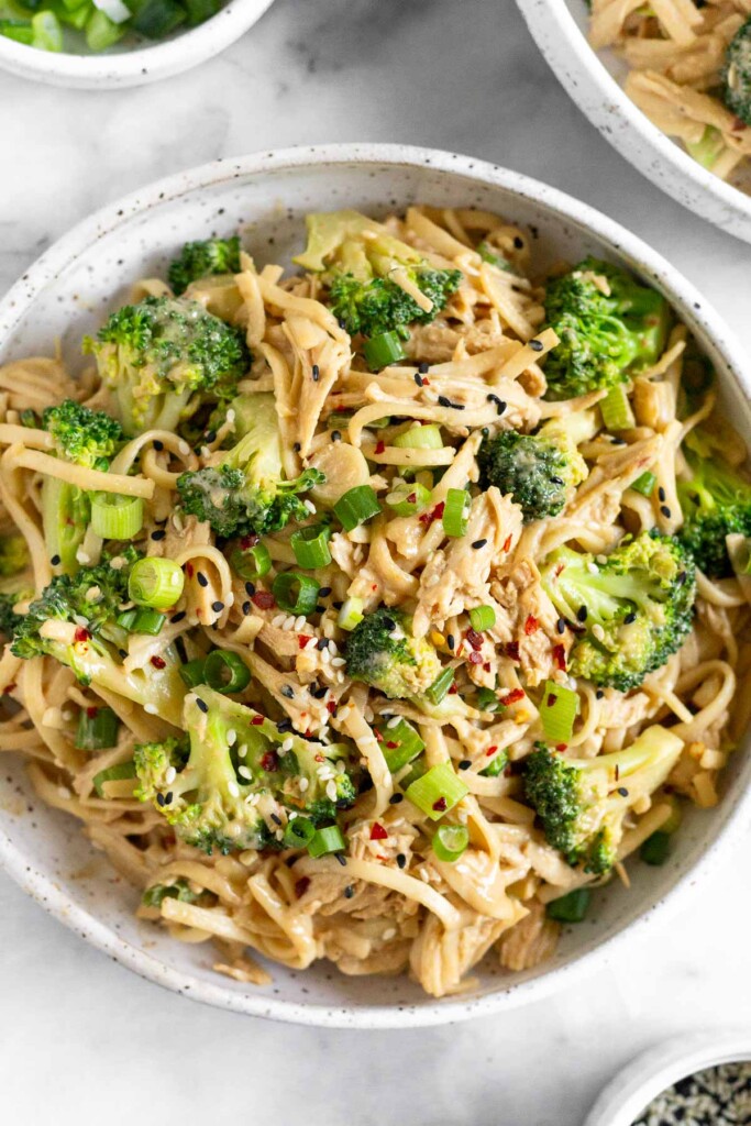 Cold sesame noodles with chicken, broccoli, and green onions in a bowl. It is garnished with sesame seeds and red pepper flakes. Around the bowl is a small dish of sesame seeds, a dish of green onions, and another bowl of cold sesame noodles.
