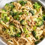 Sesame noodles with chicken and broccoli garnished with green onion, sesame seeds, and red pepper flakes in a white bowl.