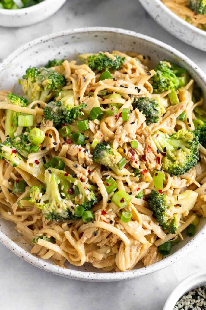 Sesame noodles with chicken and broccoli garnished with green onion, sesame seeds, and red pepper flakes in a white bowl.
