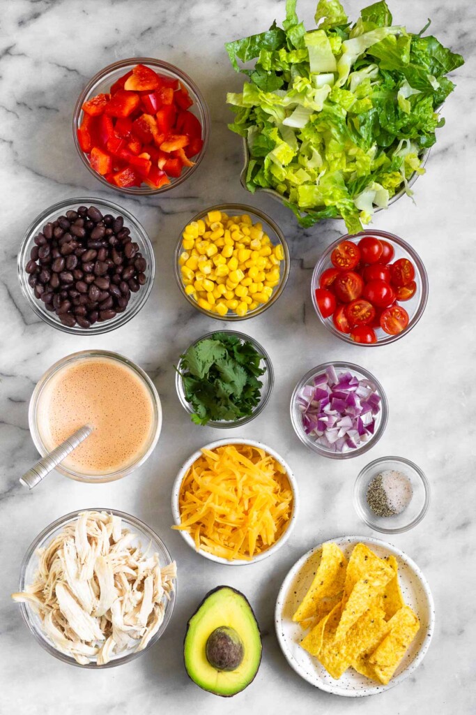 White marble counter with a bowl of chopped romaine lettuce, a bowl of diced cherry tomatoes, a bowl of diced red onion, a small bowl of salt and pepper, a plate of tortilla chips, half an avocado with the seed still in it, a bowl of shredded chicken, a bowl of shredded cheese, a jar of creamy salad dressing with a spoon in it, a bowl of cilantro, a bowl of corn, a bowl of black beans, and a bowl of diced red bell pepper.