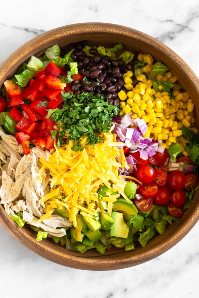 Large wooden bowl with chopped romaine, shredded chicken, diced peppers, black beans, corn, cherry tomatoes, red onion, avocado, shredded cheese, and cilantro.