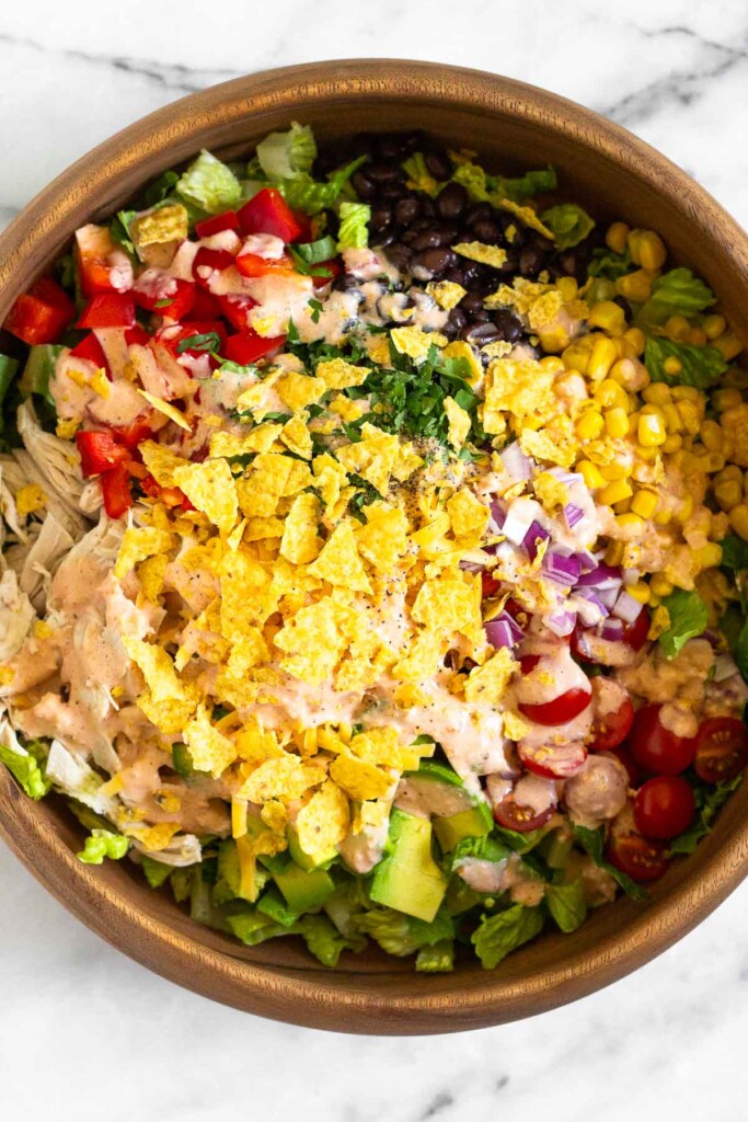 Large wooden bowl with chopped romaine, shredded chicken, diced peppers, black beans, corn, cherry tomatoes, red onion, avocado, shredded cheese, and cilantro. It is topped with a creamy salsa dressing and crushed tortilla chips.
