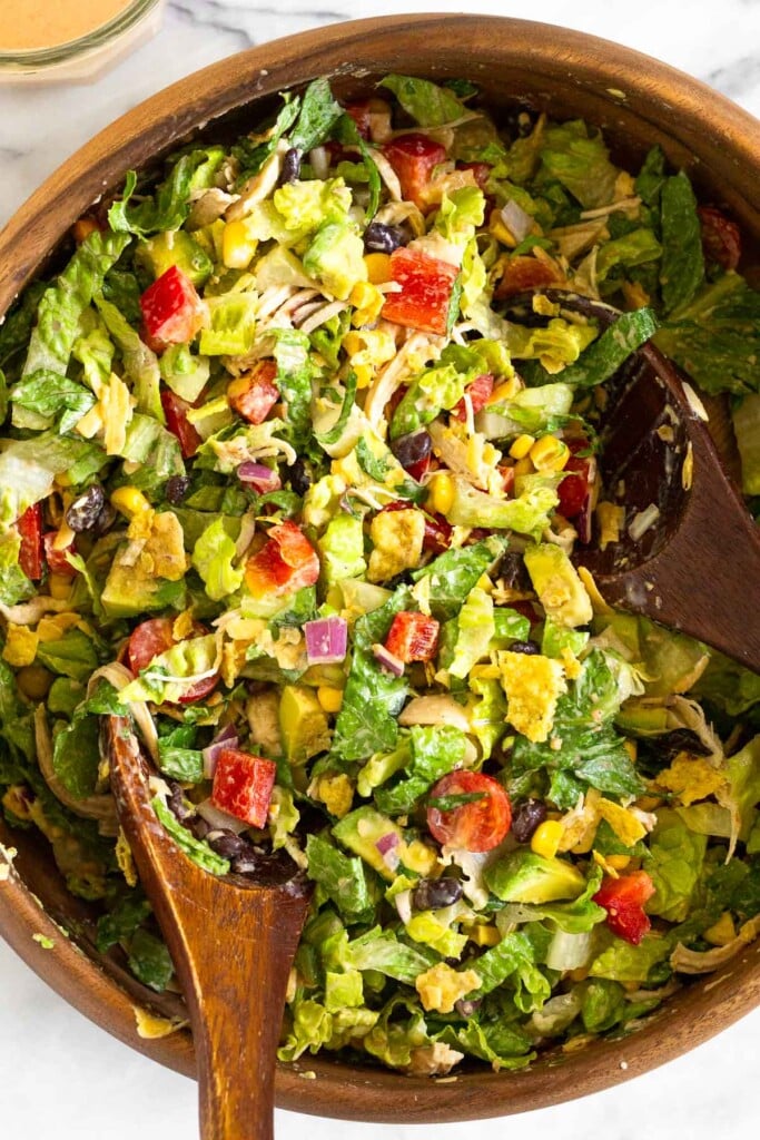 Chopped southwest chicken salad in a large wooden salad bowl with two wooden spoons in it. Off to the side of the bowl is a jar of dressing with a spoon in it.