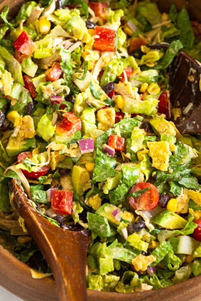 Close up of southwest chicken salad with shredded chicken, lettuce, tomatoes, peppers, beans, corn, avocado, crushed tortilla chips, and onion tossed with a salsa dressing in a large wooden bowl with two wooden spoons in it.