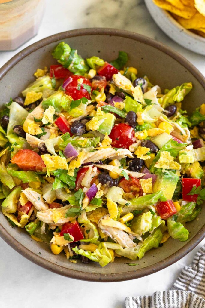 Chopped salad with lettuce, chicken, tomatoes, peppers, beans, corn, avocado, crushed tortilla chips, and onion tossed with a salsa dressing in a brown bowl. Next to the bowl is a kitchen towel, a plate of tortilla chips, and a jar of dressing.