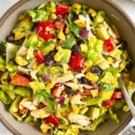 Southwest chicken salad with lettuce, tomatoes, peppers, beans, corn, avocado, crushed tortilla chips, and onion tossed with a salsa dressing in a brown bowl with a fork in it. Next to the bowl is a kitchen towel, a plate of tortilla chips, and a jar of dressing.
