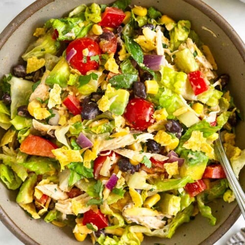 Southwest chicken salad with lettuce, tomatoes, peppers, beans, corn, avocado, crushed tortilla chips, and onion tossed with a salsa dressing in a brown bowl with a fork in it. Next to the bowl is a kitchen towel, a plate of tortilla chips, and a jar of dressing.