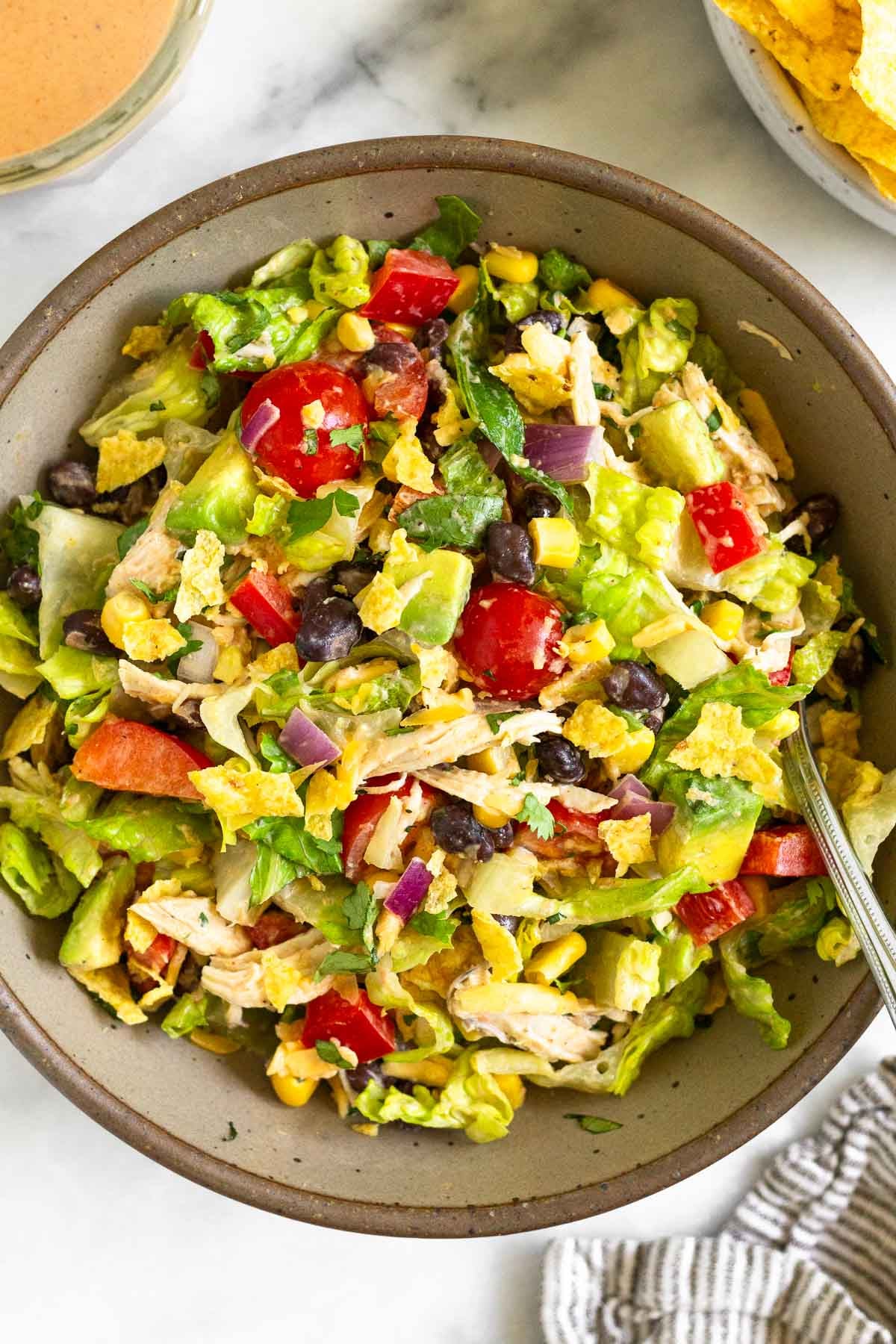 Southwest chicken salad with lettuce, tomatoes, peppers, beans, corn, avocado, crushed tortilla chips, and onion tossed with a salsa dressing in a brown bowl with a fork in it. Next to the bowl is a kitchen towel, a plate of tortilla chips, and a jar of dressing.