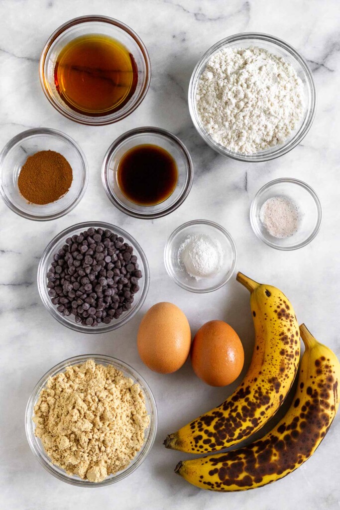 White marble counter with a bowl of flour, a small bowl of salt, a bowl of baking powder, two brown eggs, two spotted bananas, a bowl of protein powder, a bowl of mini chocolate chips, a bowl of vanilla extract, a bowl of cinnamon, and a bowl of maple syrup.