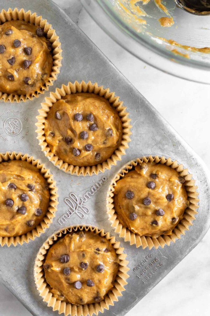 Muffin tin filled with uncooked muffin batter with chocolate chips in it. Off to the side is a empty  large bowl with a little batter in it.