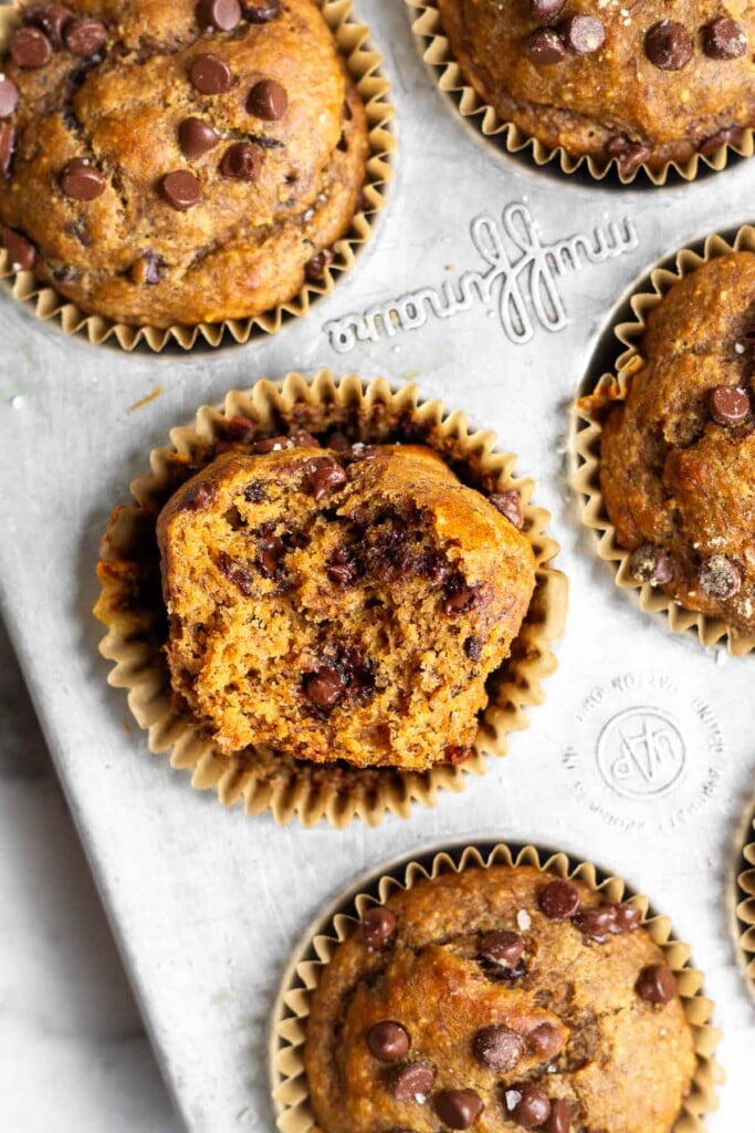 An overhead shot of chocolate chip protein muffins in a muffin tin. One of the muffins is turned on its side with a bite taken out of it.