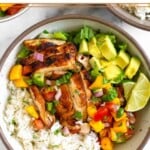 Chicken and rice bowl with coconut rice, grilled chicken thighs, romaine, fresh mango cucumber salsa, diced avocado, and two limes wedges.