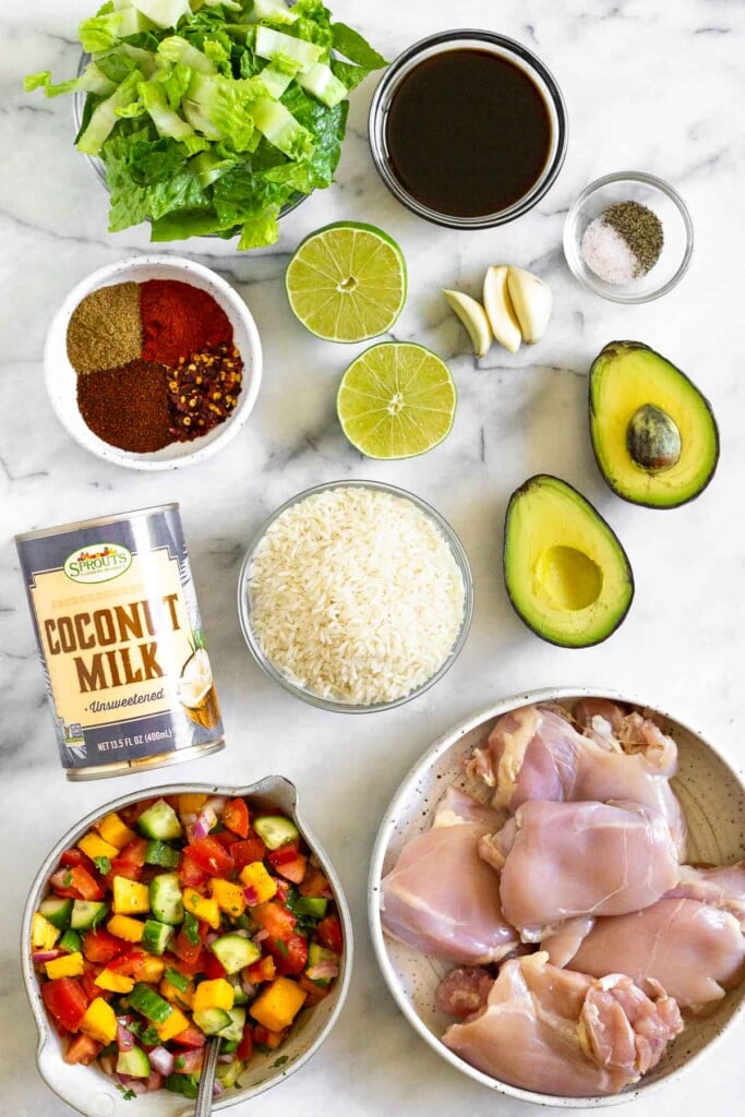 White marble counter with a bowl of coconut aminos, a small bowl of salt and pepper, 3 cloves of garlic, an avocado cut in half, a bowl of raw chicken thighs, a bowl of fresh mango cucumber salsa, a bowl of uncooked white rice, a can of coconut milk, a bowl of spices, a lime cut in half, and a bowl of chopped romaine lettuce.