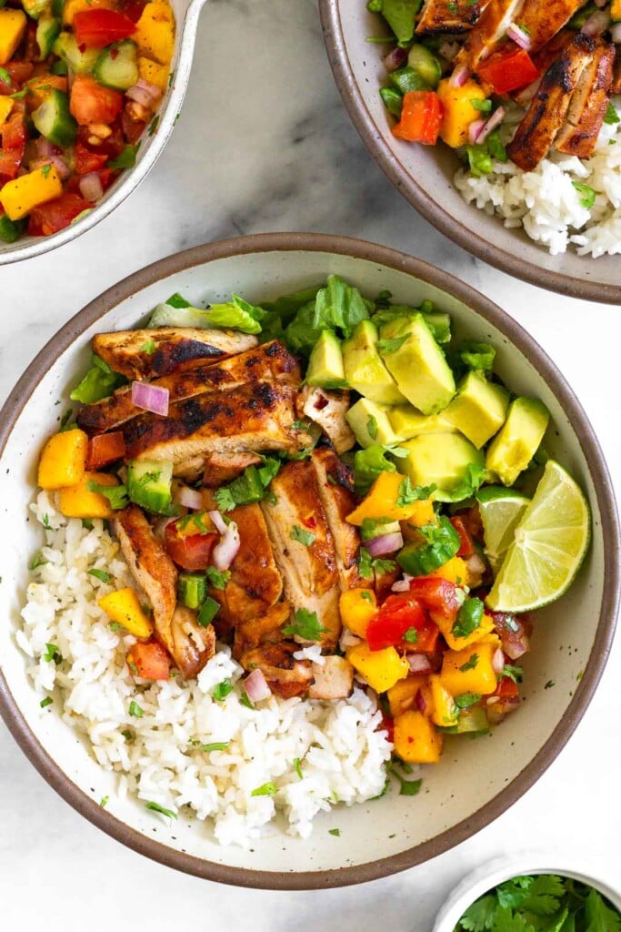 Overhead shot of a grilled chicken bowl with coconut rice, romaine, mango cucumber salsa, avocado, and two lime wedges. Around the bowl is a small dish of cilantro, another grilled chicken bowl, and a bowl of mango cucumber salsa.