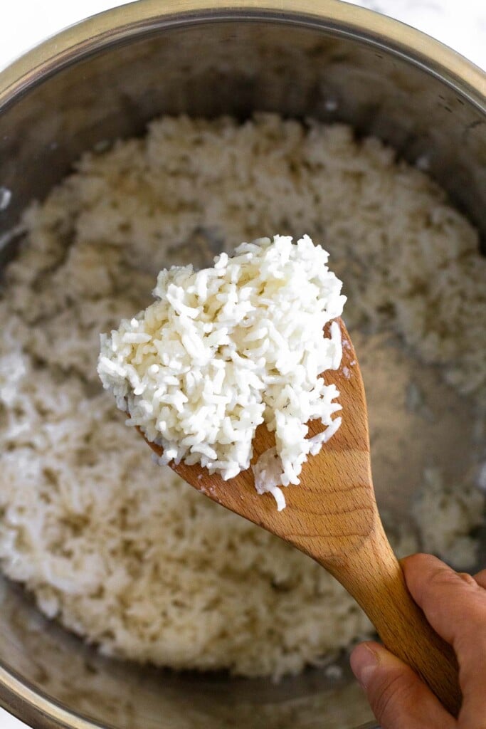 Overhead shot of scoop of coconut rice on a wooden spoon. Underneath it is a pot with more rice in it.