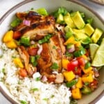 Chicken and rice bowl with coconut rice, grilled chicken thighs, romaine, fresh mango cucumber salsa, diced avocado, and two limes wedges. Around the bowl is a small dish of cilantro, another chicken and rice bowl, and a bowl of mango salsa.