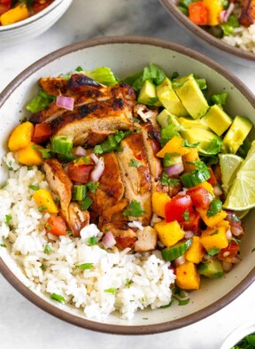 Chicken and rice bowl with coconut rice, grilled chicken thighs, romaine, fresh mango cucumber salsa, diced avocado, and two limes wedges. Around the bowl is a small dish of cilantro, another chicken and rice bowl, and a bowl of mango salsa.