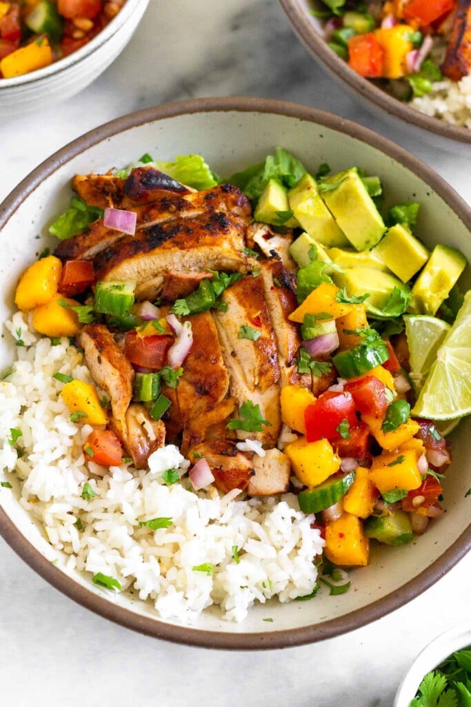 Chicken and rice bowl with coconut rice, grilled chicken thighs, romaine, fresh mango cucumber salsa, diced avocado, and two limes wedges. Around the bowl is a small dish of cilantro, another chicken and rice bowl, and a bowl of mango salsa.