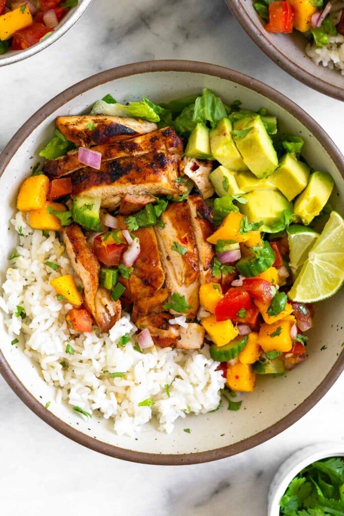 Overhead shot of a white bowl with coconut rice, grilled chicken thighs, romaine, mango salsa, avocado, and lime wedges. Around the bowl is a small dish of cilantro, another bowl with rice and salsa, and a bowl of mango salsa.