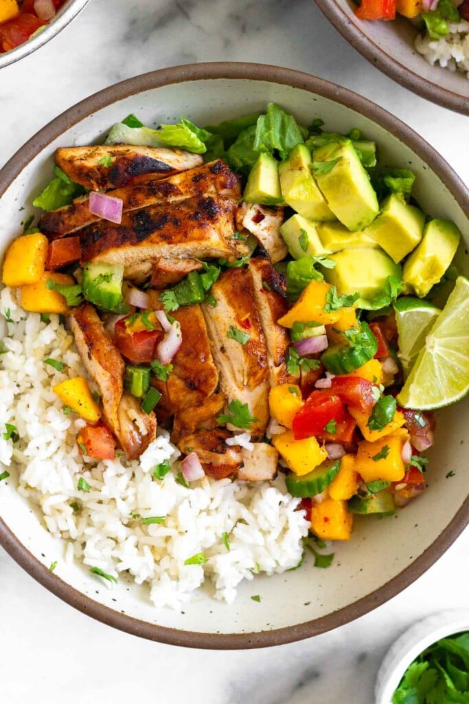 Chicken rice bowl with coconut rice, grilled chicken thighs, mango salsa, romaine, avocado, and 2 lime wedges.