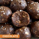 A close up of a pile of chocolate peanut butter protein balls sprinkled with flaky sea salt. The one in the front as a bite taken out of it.